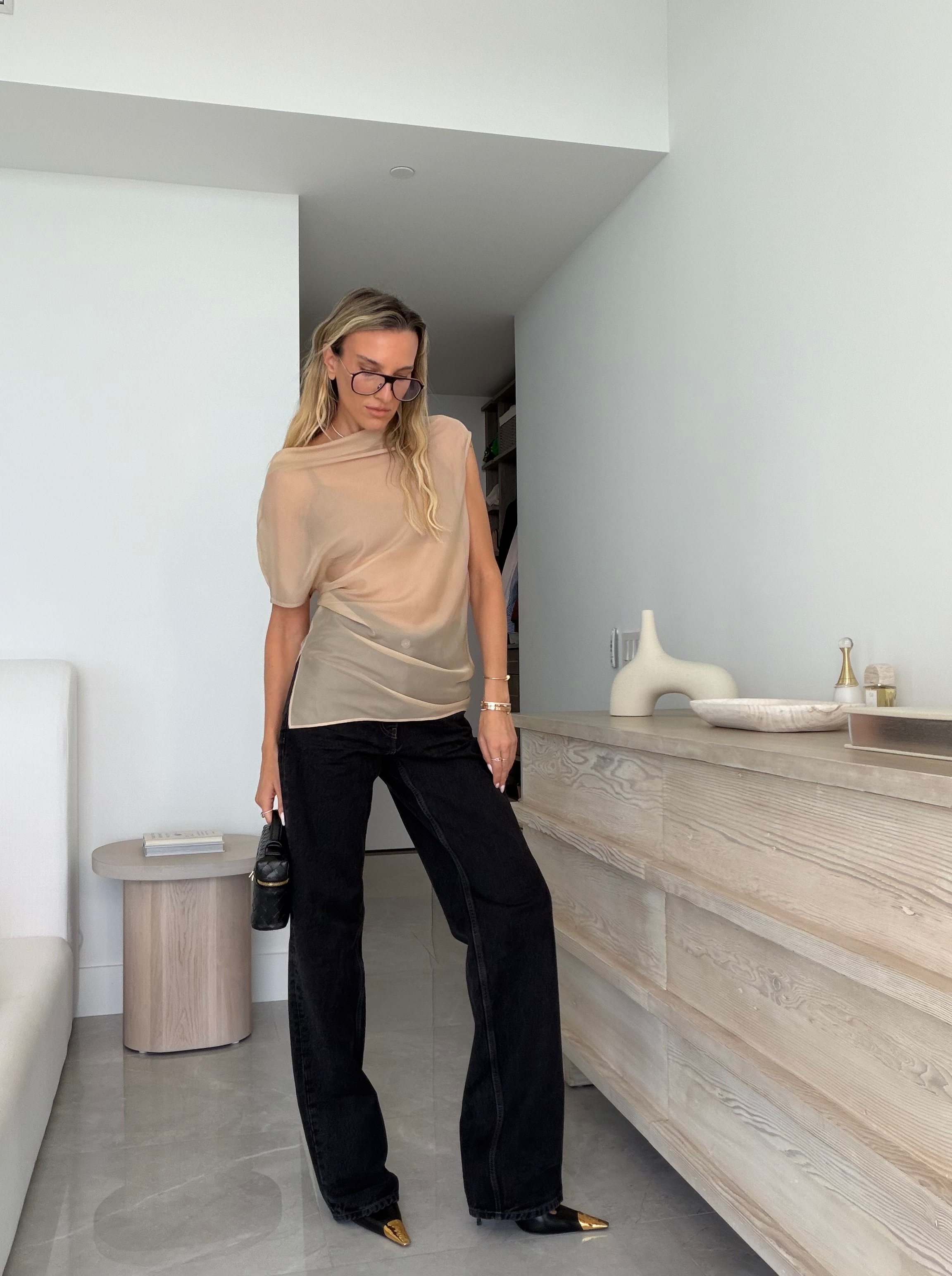 Woman wearing sheer beige Eaves top from Revolve styled with Saint Laurent black v-waist jeans and pointed toe heels, modern daytime outfit