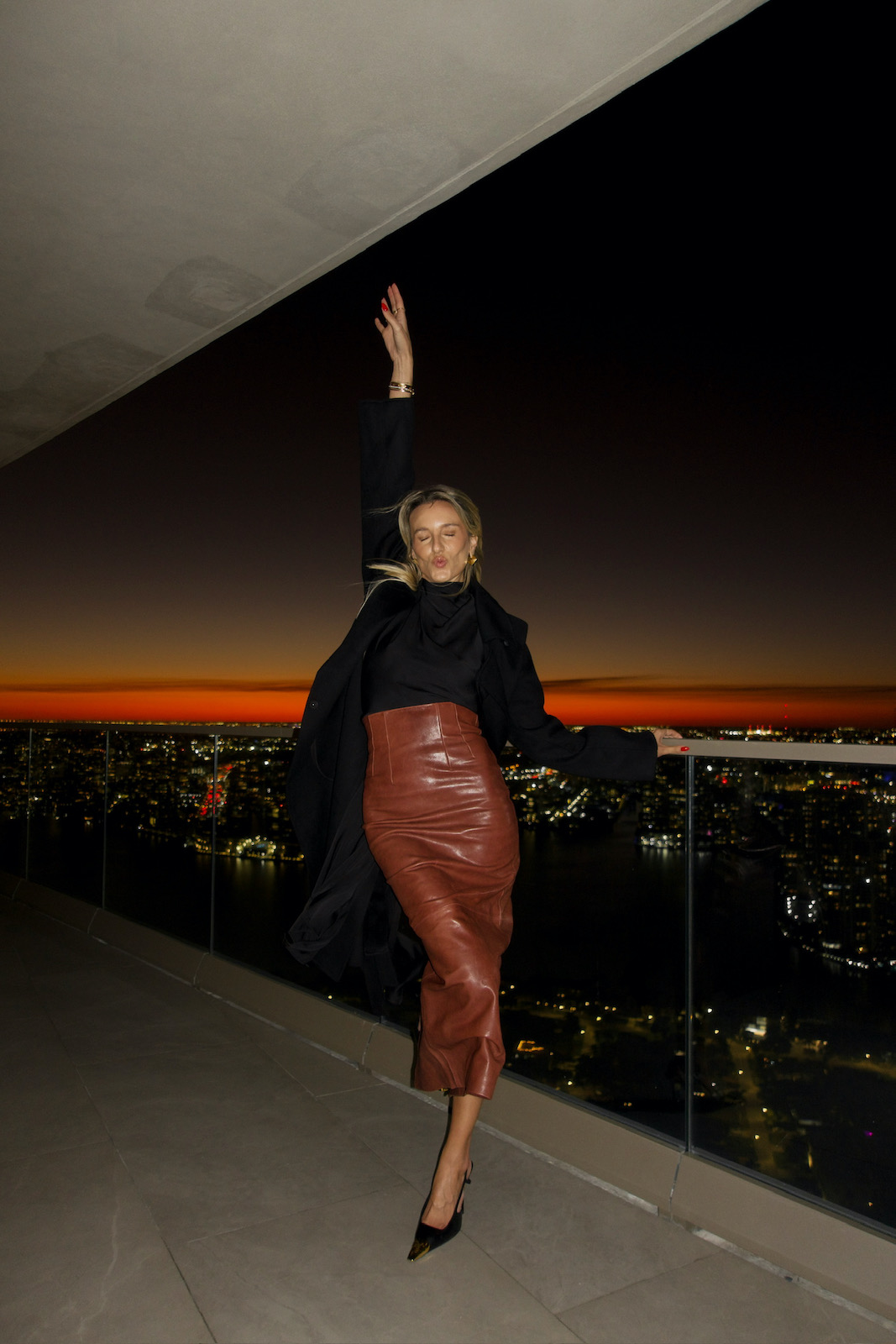 Woman styling a brown leather skirt with a black top, cashmere coat, and heels against a city skyline at night.