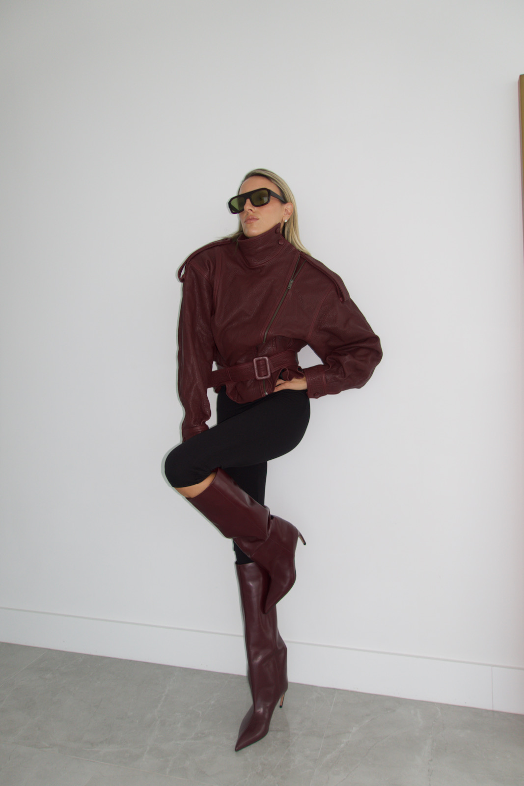 Woman wearing a burgundy leather jacket styled with black leggings and knee-high burgundy boots in a minimalist interior